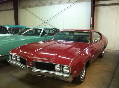 Red vintage muscle car displayed in indoor garage with green car visible in background
