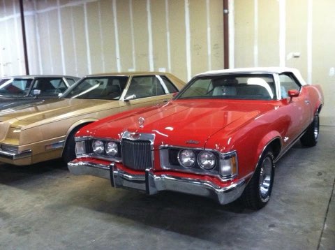 Red 1970s Mercury Cougar convertible displayed in showroom with tan sedan