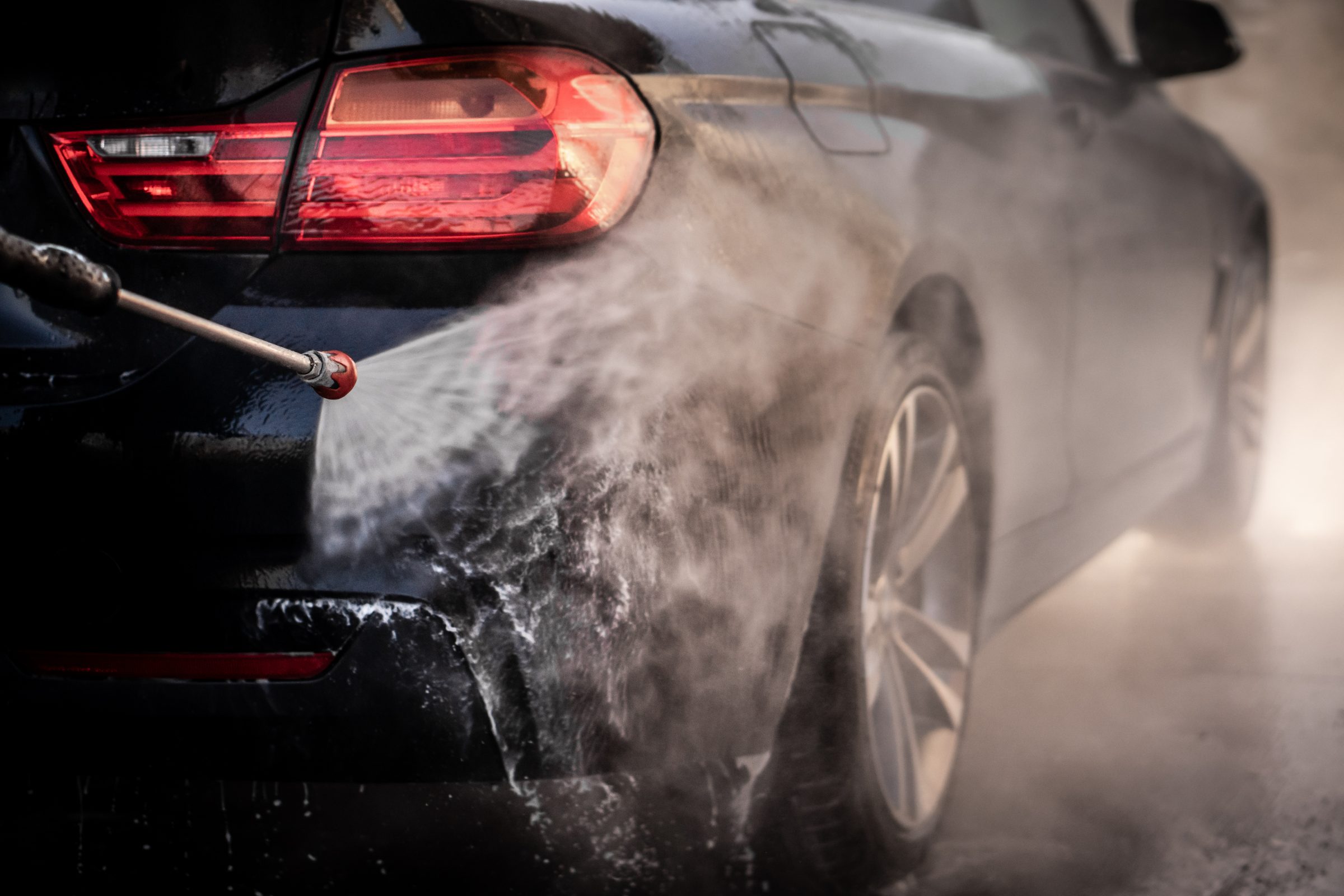 High-pressure water spray cleaning the side of a dark car during professional washing service